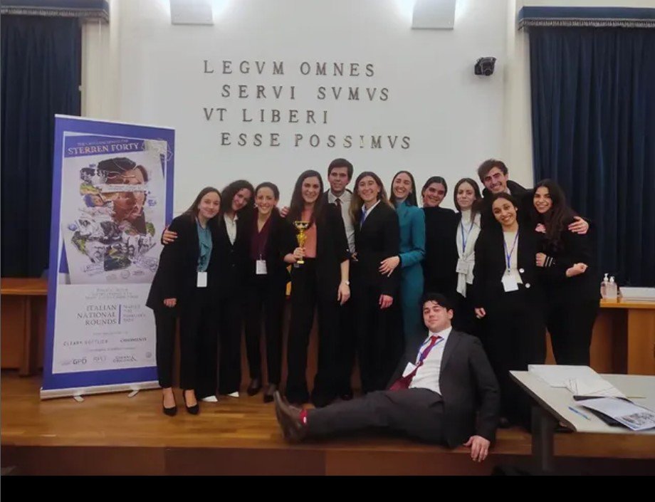 A group of young people dressed in formal attire, standing on a stage, holding a trophy, with one person sitting in front. There is a banner to the left and Latin text on the wall behind them.
