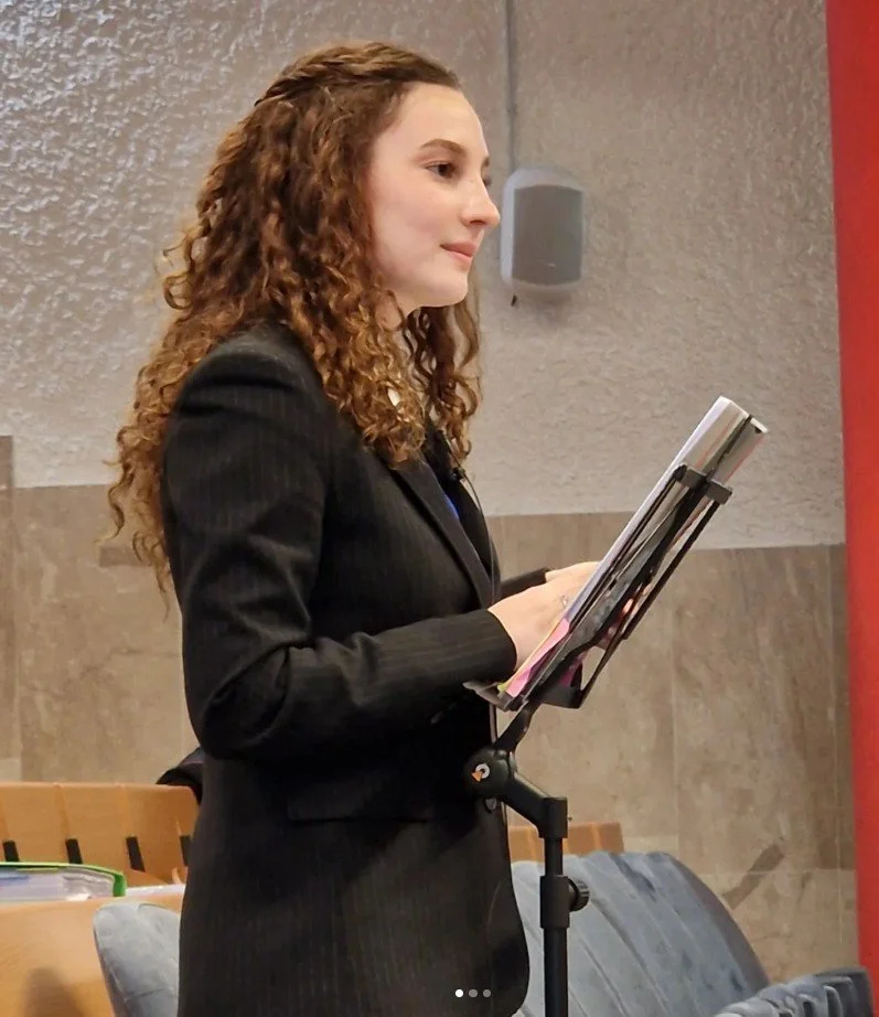 A young woman with curly brown hair wearing a black blazer standing at a music stand, reading or preparing to speak, in a formal setting with beige and red walls.