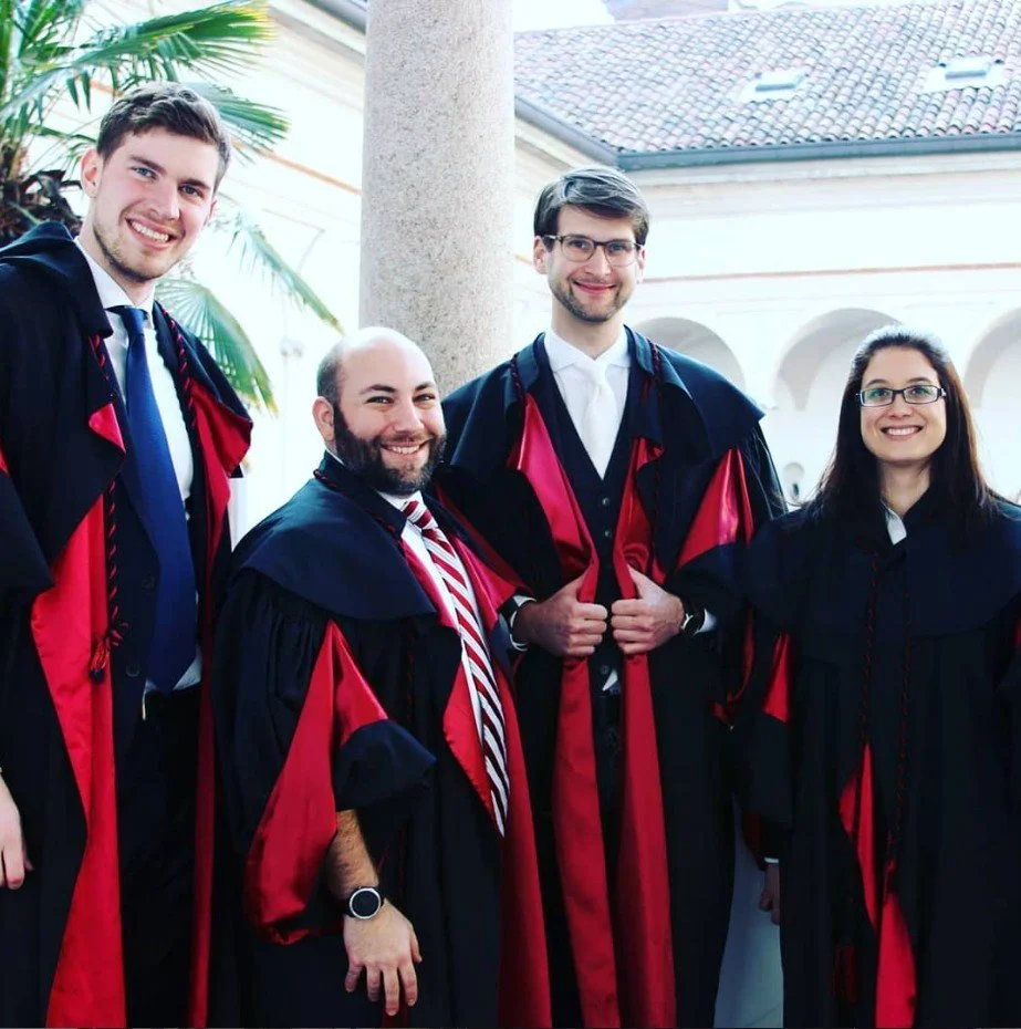 Four people in academic regalia posing for a photo during a graduation ceremony, with a column and a building with arches in the background.
