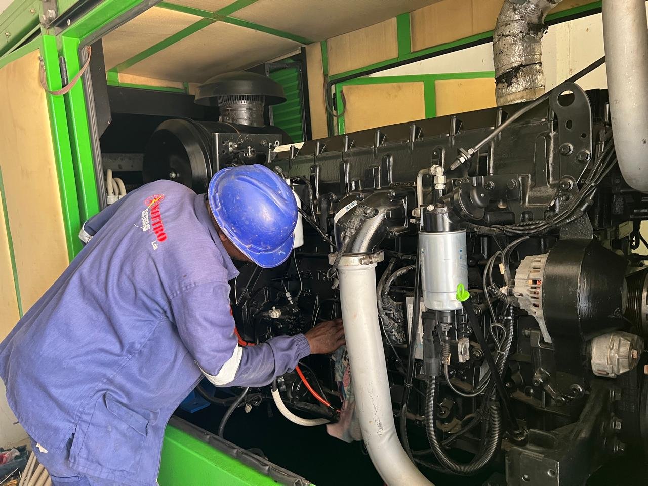 Um técnico usando capacete azul trabalha na manutenção de um motor de grande porte, instalado em um equipamento com estrutura de metal verde.