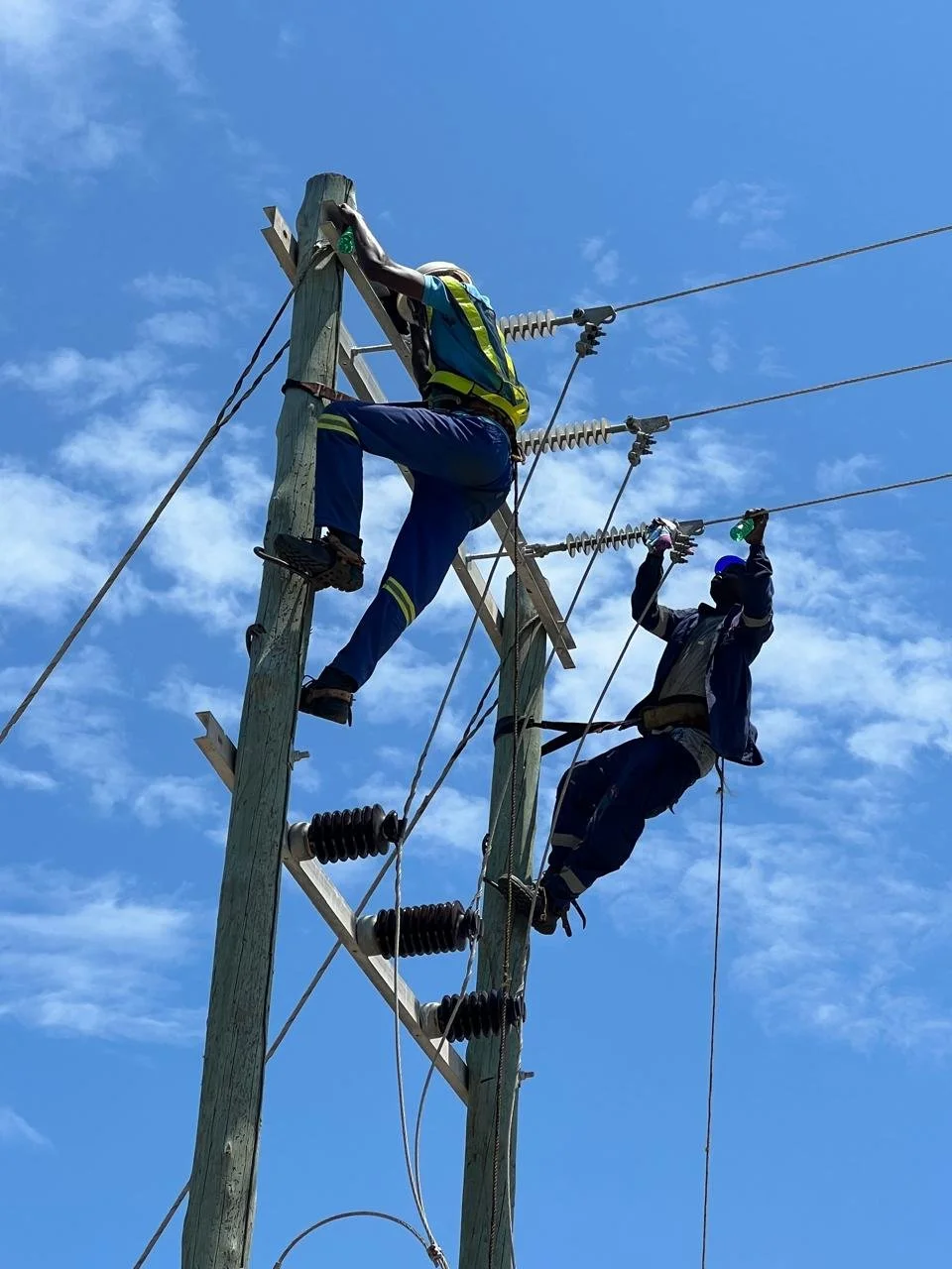 Dois técnicos trabalhando na manutenção de um poste de energia elétrica ao ar livre durante o dia.