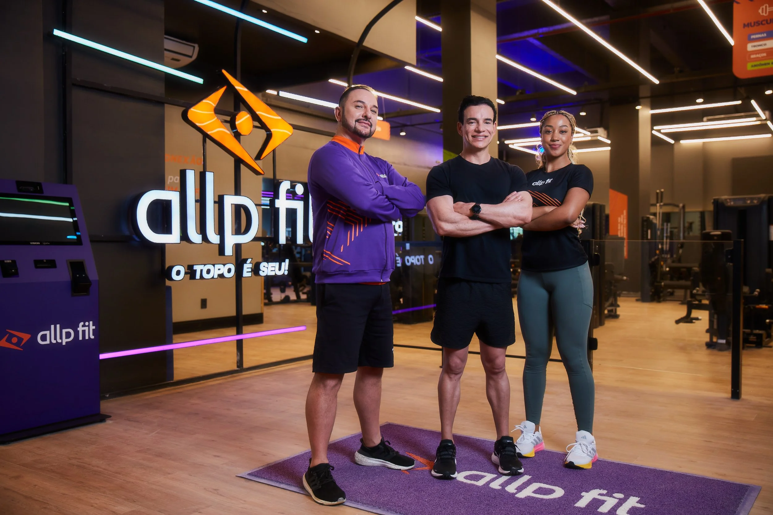 Three people standing on a purple workout mat in a gym, smiling at the camera, with gym equipment visible in the background. Two men and one woman, dressed in athletic wear, pose with arms crossed.