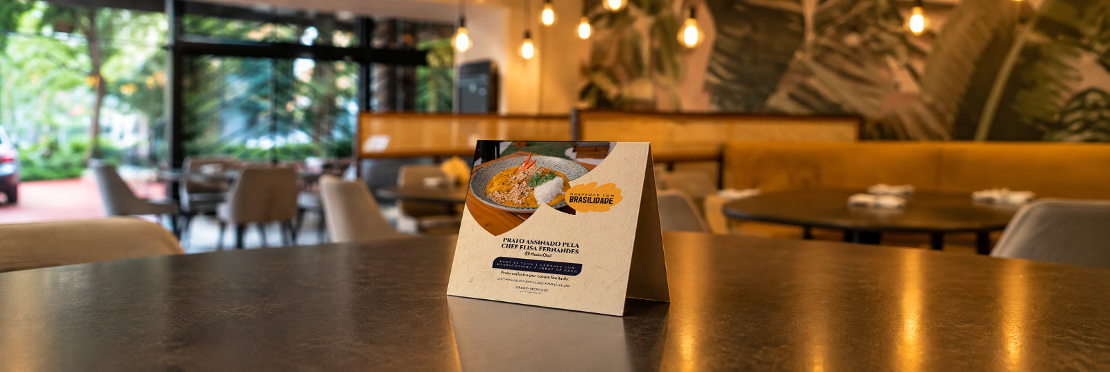 A table tent card featuring an image of a plated dish, displayed on a dining table inside a restaurant with large windows, tables, chairs, warm lighting, and tropical decor.