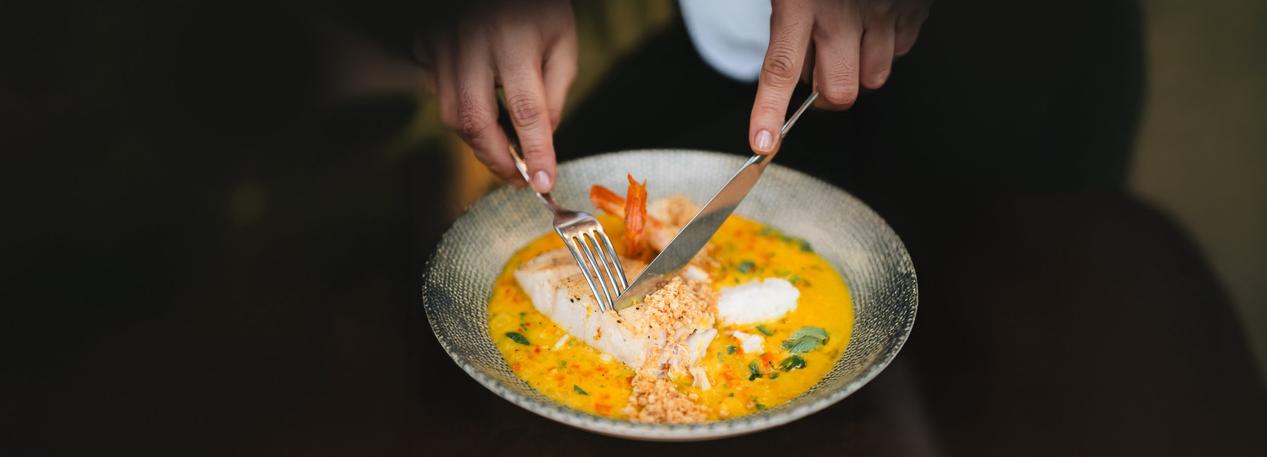 Person cutting fish fillet on a plate of yellow sauce with herbs and breadcrumbs.