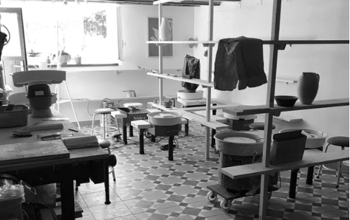 Ein Kunststudio mit Tischen, Regalen, Stoffen und Kunstmaterialien in einem Raum mit Fenstern.