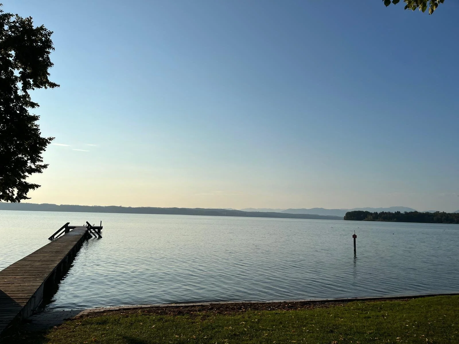 Blick auf einen See bei Sonnenuntergang, mit einem Steg im Wasser und einem Pfosten im Wasser, umgeben von Bäumen und Hügeln im Hintergrund.