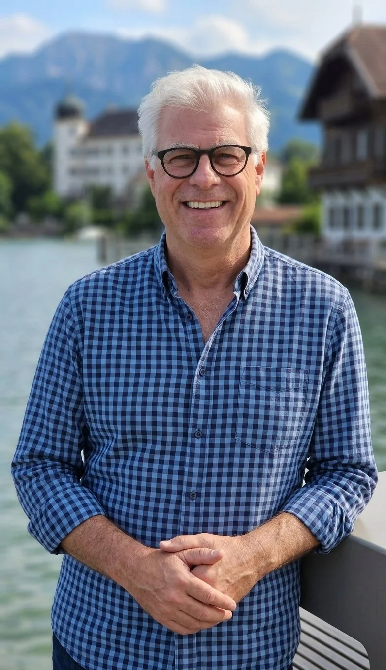 Ein lächelnder älterer Mann mit weißen Haaren und Brille, trägt ein blau kariertes Hemd, steht draußen an einem Wasser mit Bergen im Hintergrund.