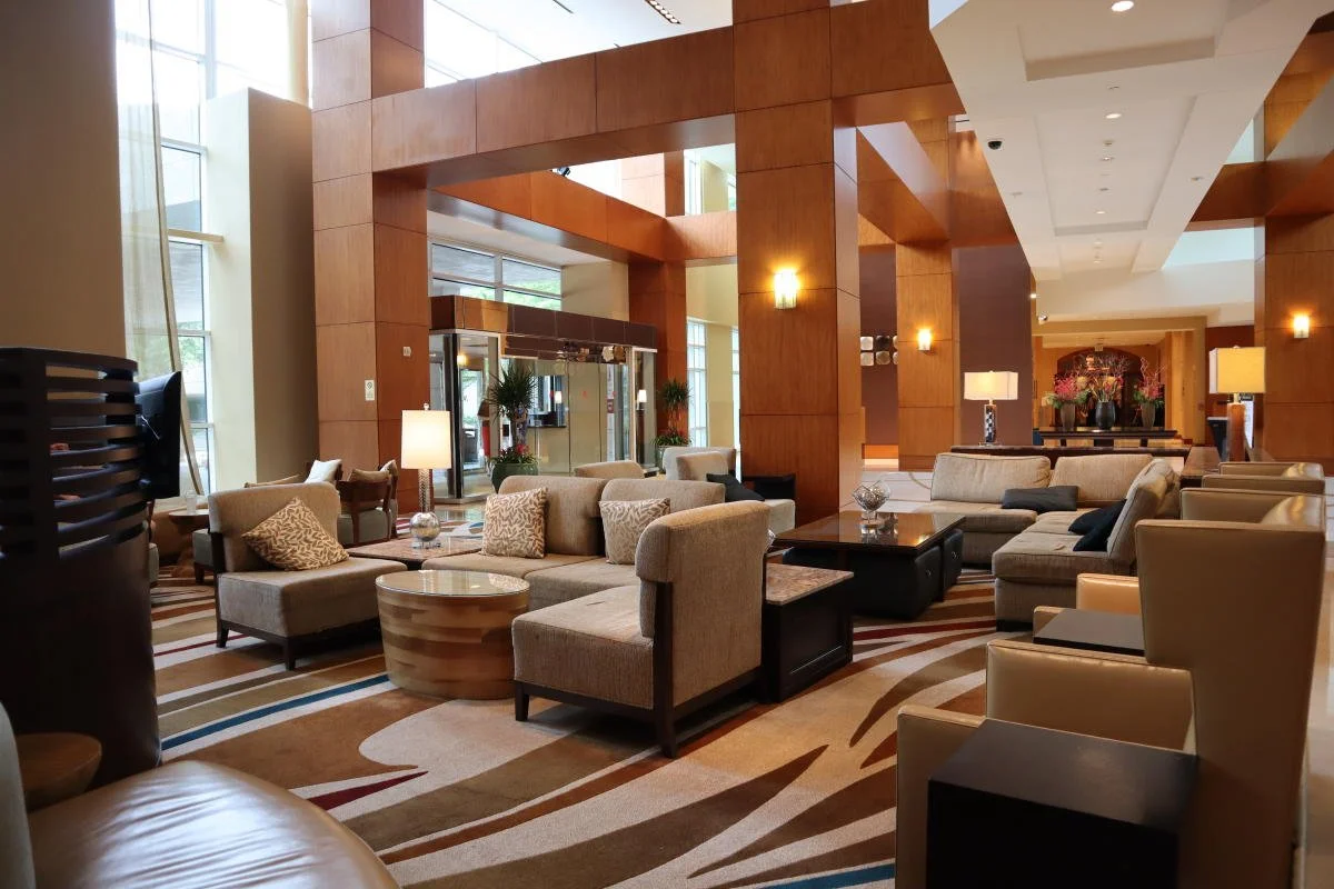 Hotel lobby with beige and brown sofas, armchairs, coffee tables, and decorative lamps, featuring large windows and wooden paneling.