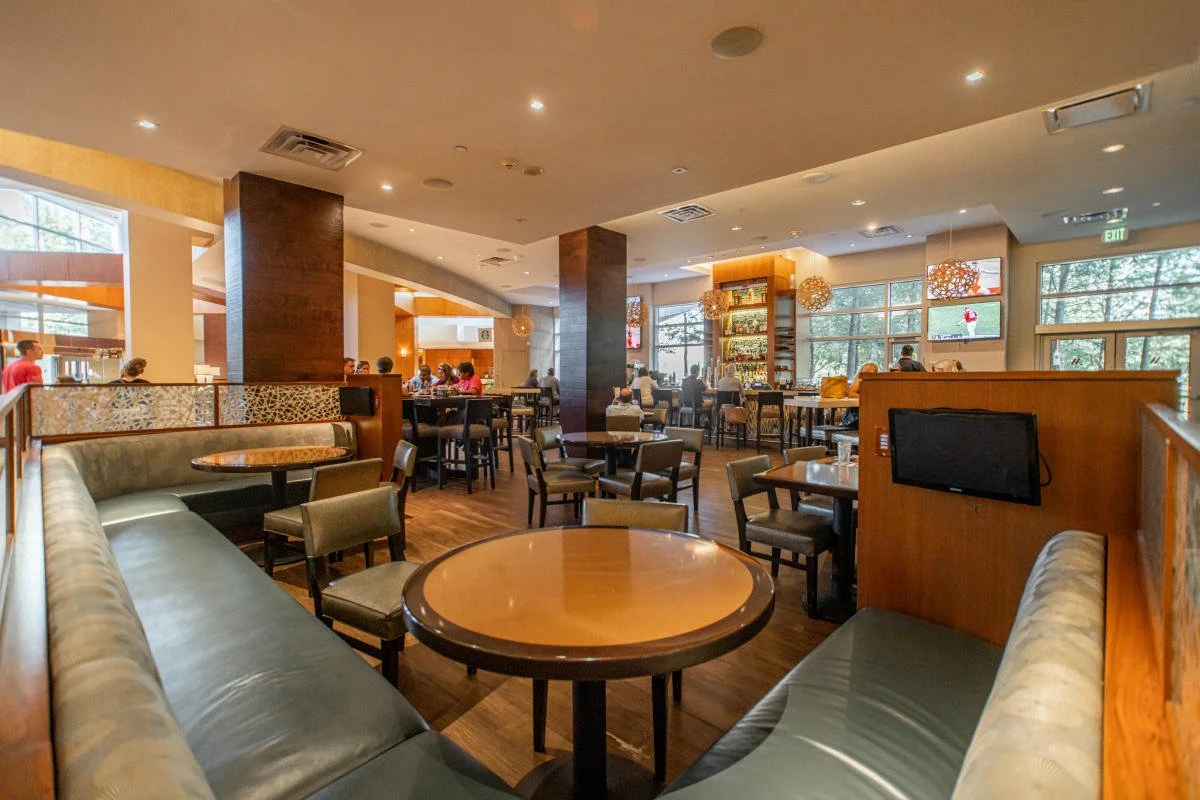 Interior view of a restaurant with multiple tables and chairs, some patrons seated, large windows allowing natural light, decorative hanging lamps, a TV on the wall, and a bar area in the background.