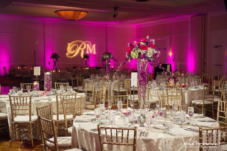 Elegant banquet hall decorated for a wedding reception with round tables covered in white tablecloths, gold chairs, floral centerpieces, and pink uplighting on the walls. A monogrammed projection 'DR' is visible on the wall in the background.