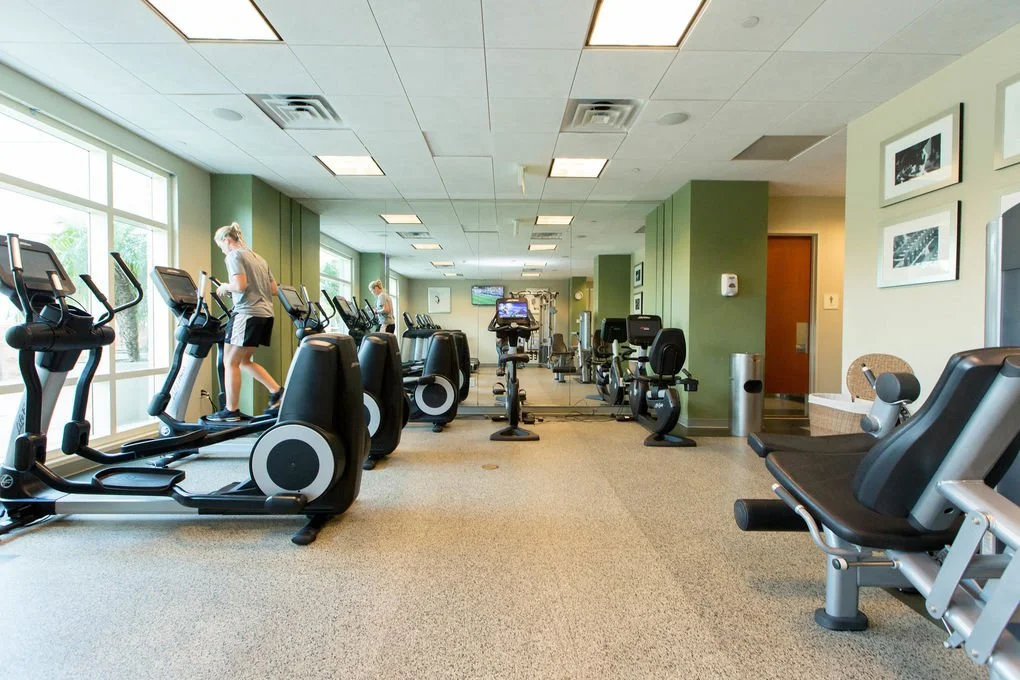 Fitness center with cardio equipment, including treadmills and stationary bikes, with people exercising, large windows, and framed pictures on the wall.