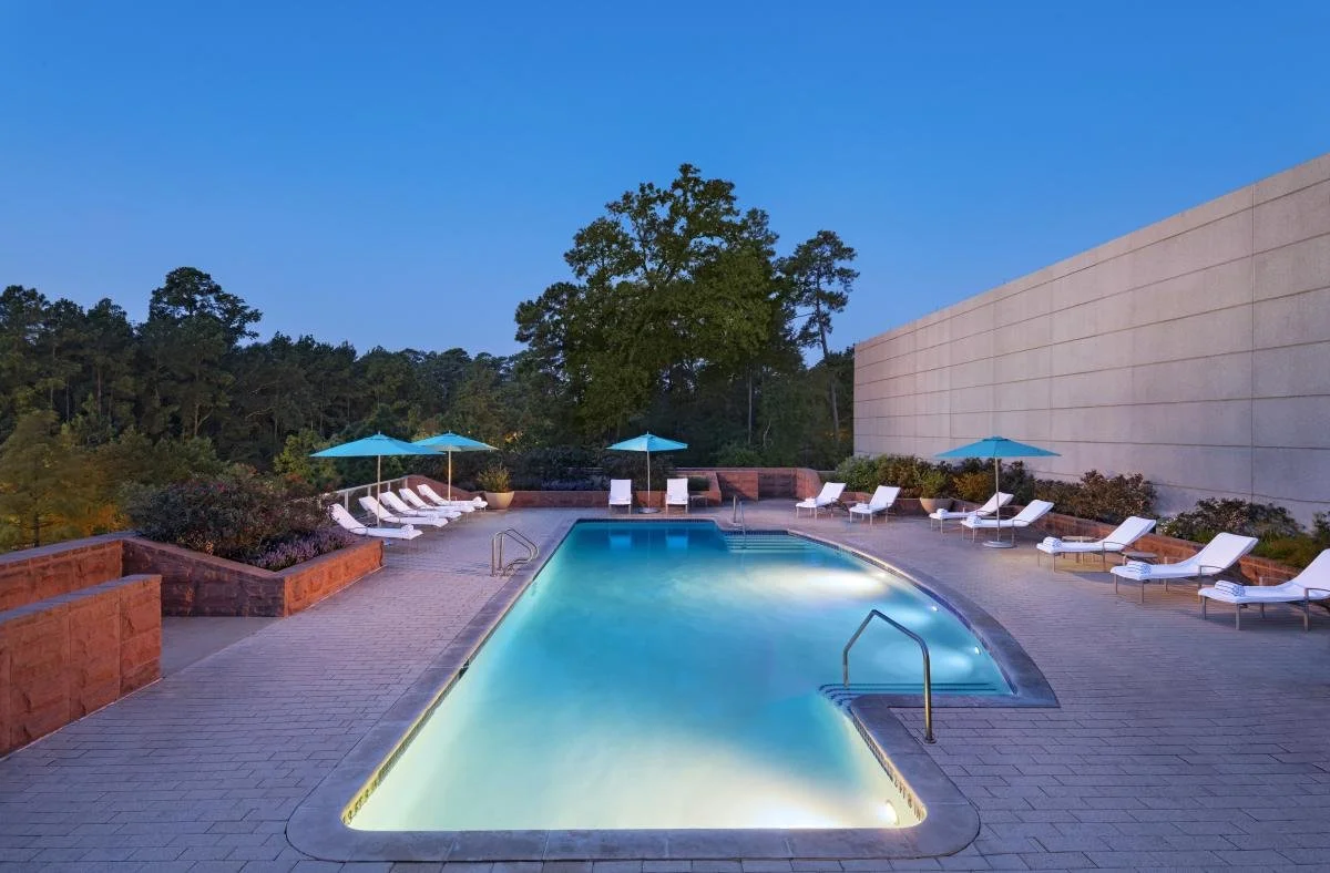 Outdoor swimming pool surrounded by lounge chairs with white cushions and blue umbrellas at dusk, with trees and a brick wall in the background.
