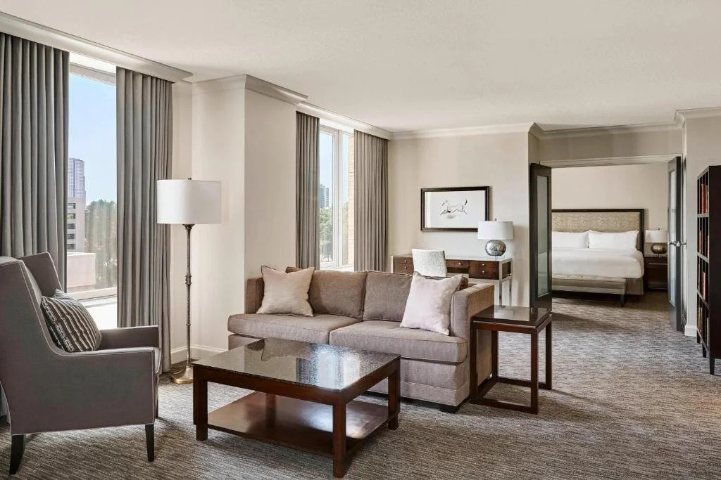 Hotel suite with a seating area, large windows with curtains, a bed in a separate room, and modern furniture.