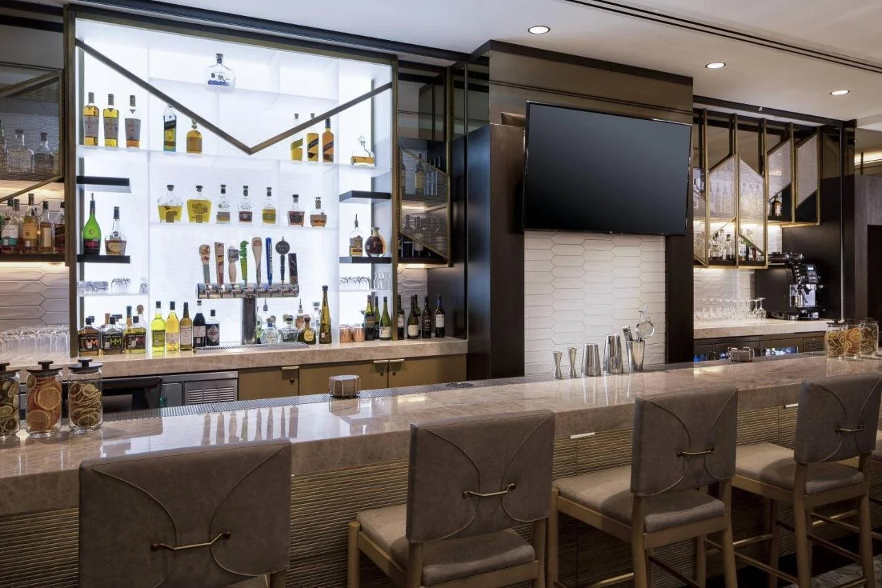 A modern bar with a marble countertop, cushioned bar stools, and a backlit liquor display with various bottles and glasses, along with a large TV mounted on the wall.