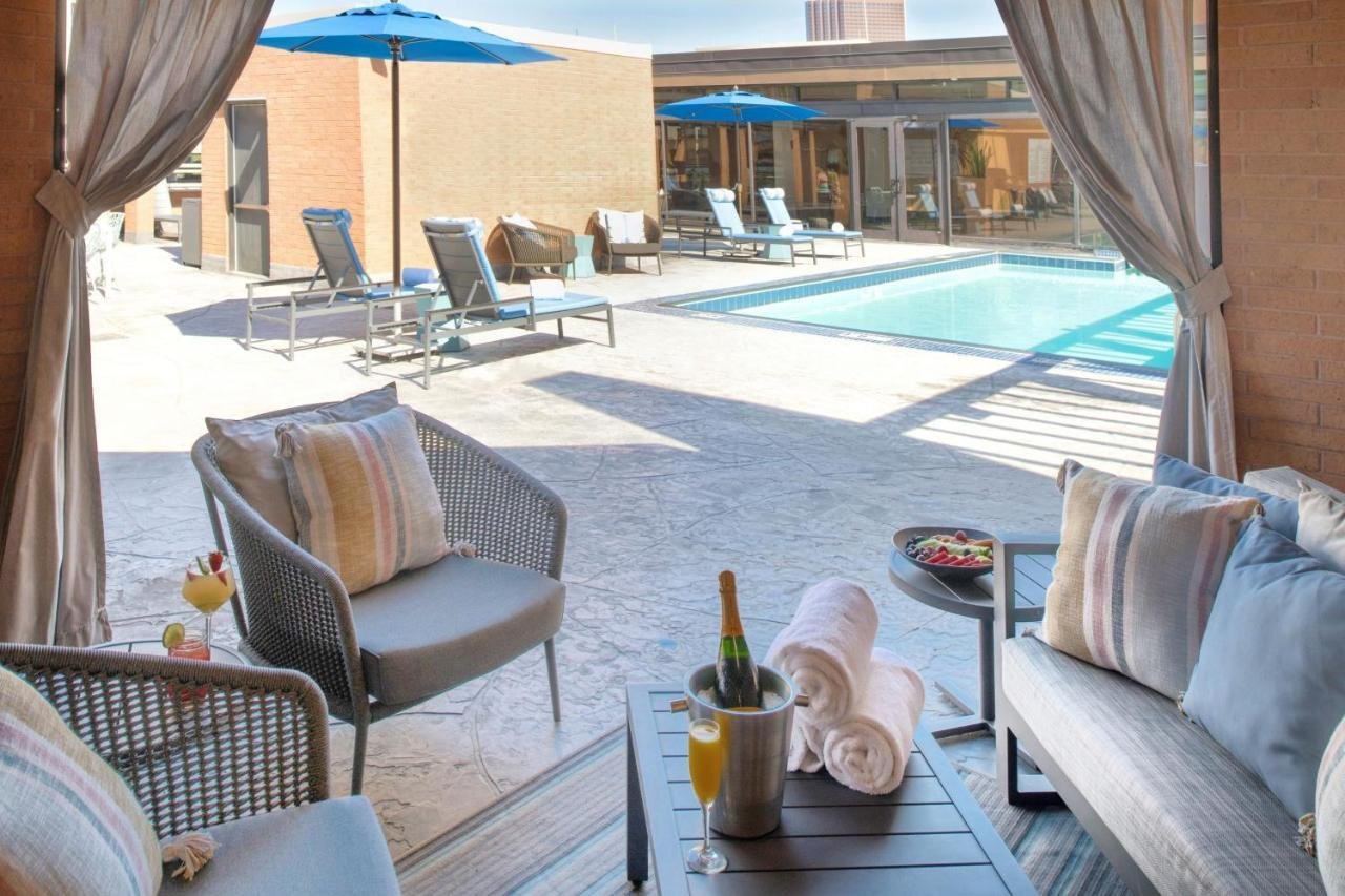 Outdoor pool area viewed from a covered patio with seating, towels, a bottle of champagne, and glasses, with lounge chairs and umbrellas around the pool.