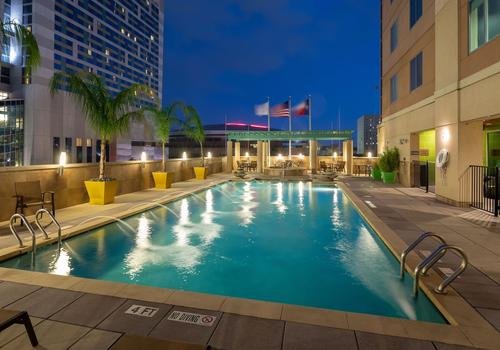Rooftop swimming pool at dusk with palm trees, lounge chairs, and flags in the background