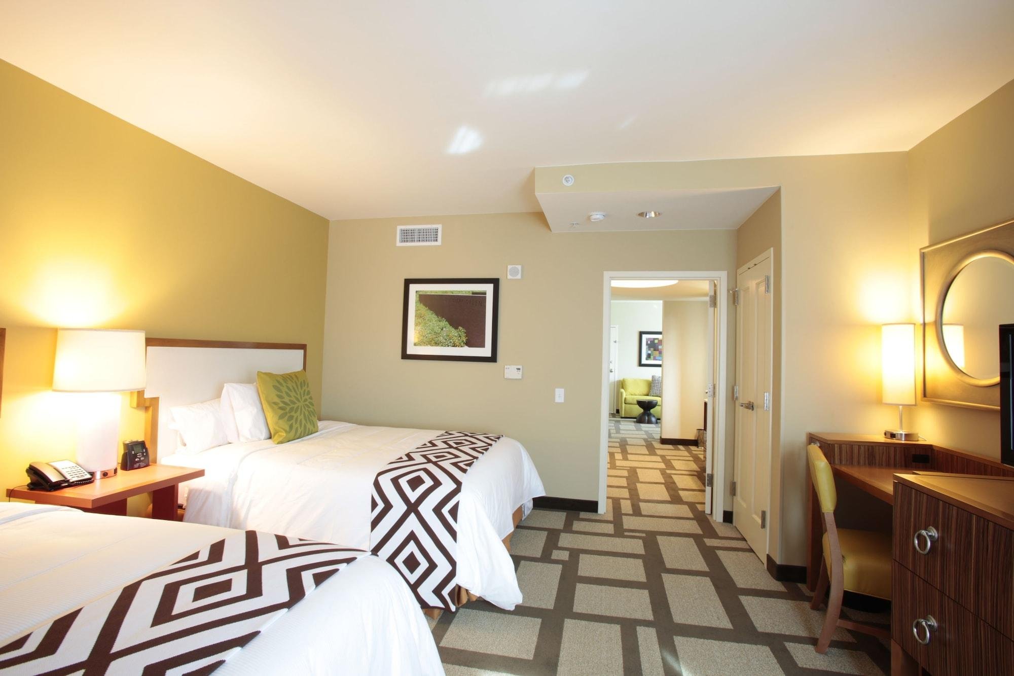 A hotel room with two beds, bedside lamps, a framed picture on the wall, a window in the doorway leading to a sitting area, a desk with a chair, and patterned carpet.