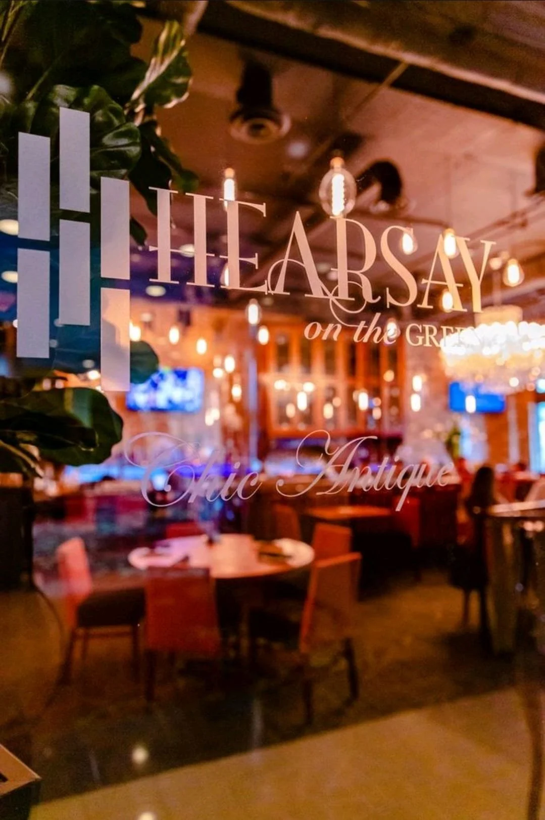 View of a restaurant interior with wooden furniture, hanging Edison bulbs, and a large mirror or glass window with writing on it that says 'The Arsay on the Great' and 'Nuc Antique.'