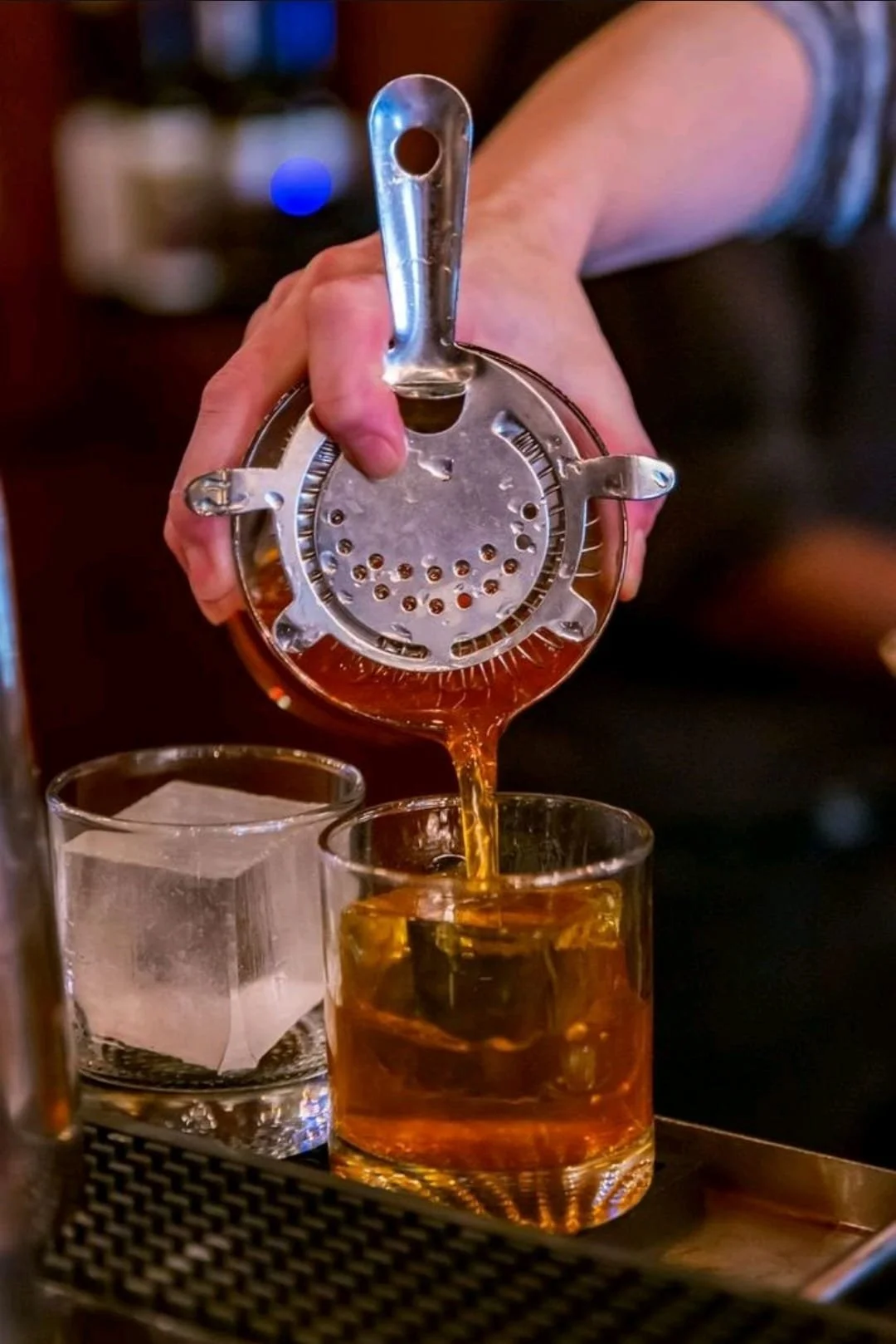 Person pouring whiskey through a metal strainer into a glass, with a large ice cube nearby.