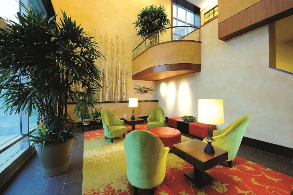 Hotel lobby with yellow walls, large green potted plant, bright green armchairs, red ottomans, side tables with lamps, and a staircase with a glass railing.