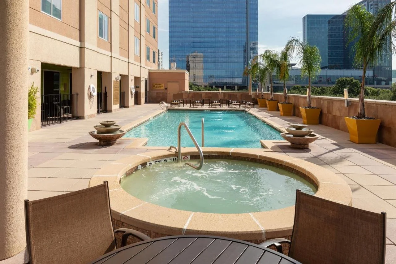 Rooftop pool and hot tub in an urban setting, with tall skyscrapers, lounge chairs, and potted palm trees.
