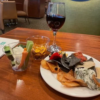 A glass of red wine, a plate with assorted cheeses, crackers, and vegetables, and a small bowl of mixed nuts on a wooden table in a restaurant setting.