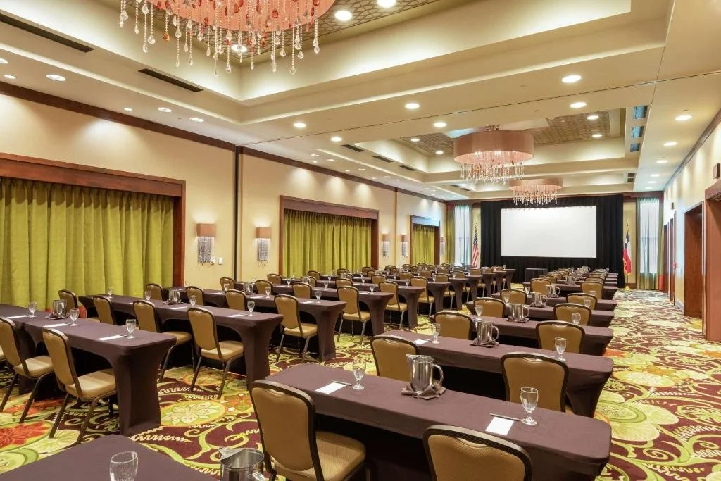 A large conference room set up with rows of tables and chairs, each table has water glasses and pitchers, a stage with a screen, chandeliers hanging from the ceiling, and green curtains covering the windows.