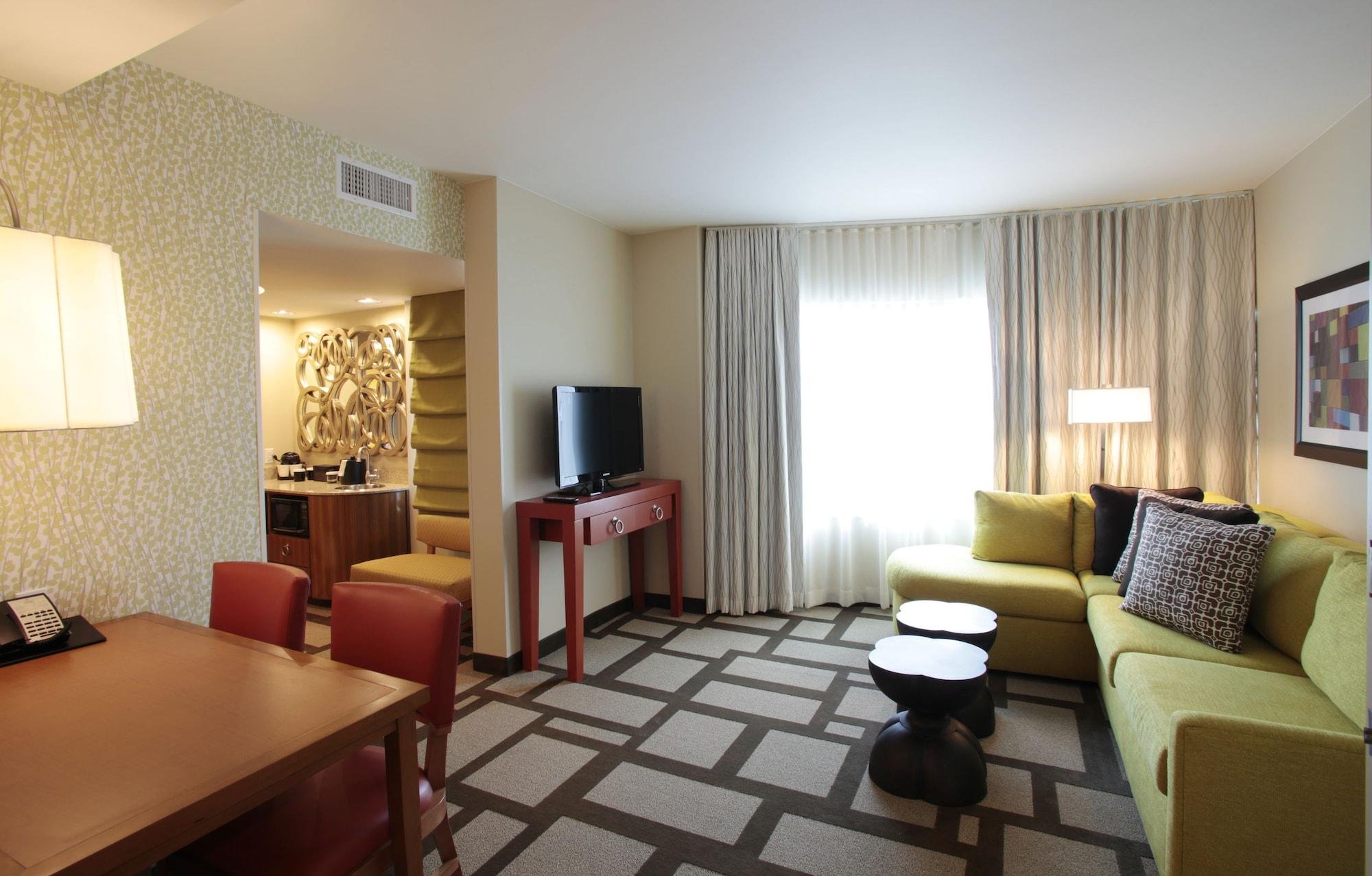 L-shaped green sofa with patterned pillows, small round black tables, window with beige curtains, flat-screen TV on red stand, part of a dining table with red chairs, patterned carpet, lamp, and artwork on wall.