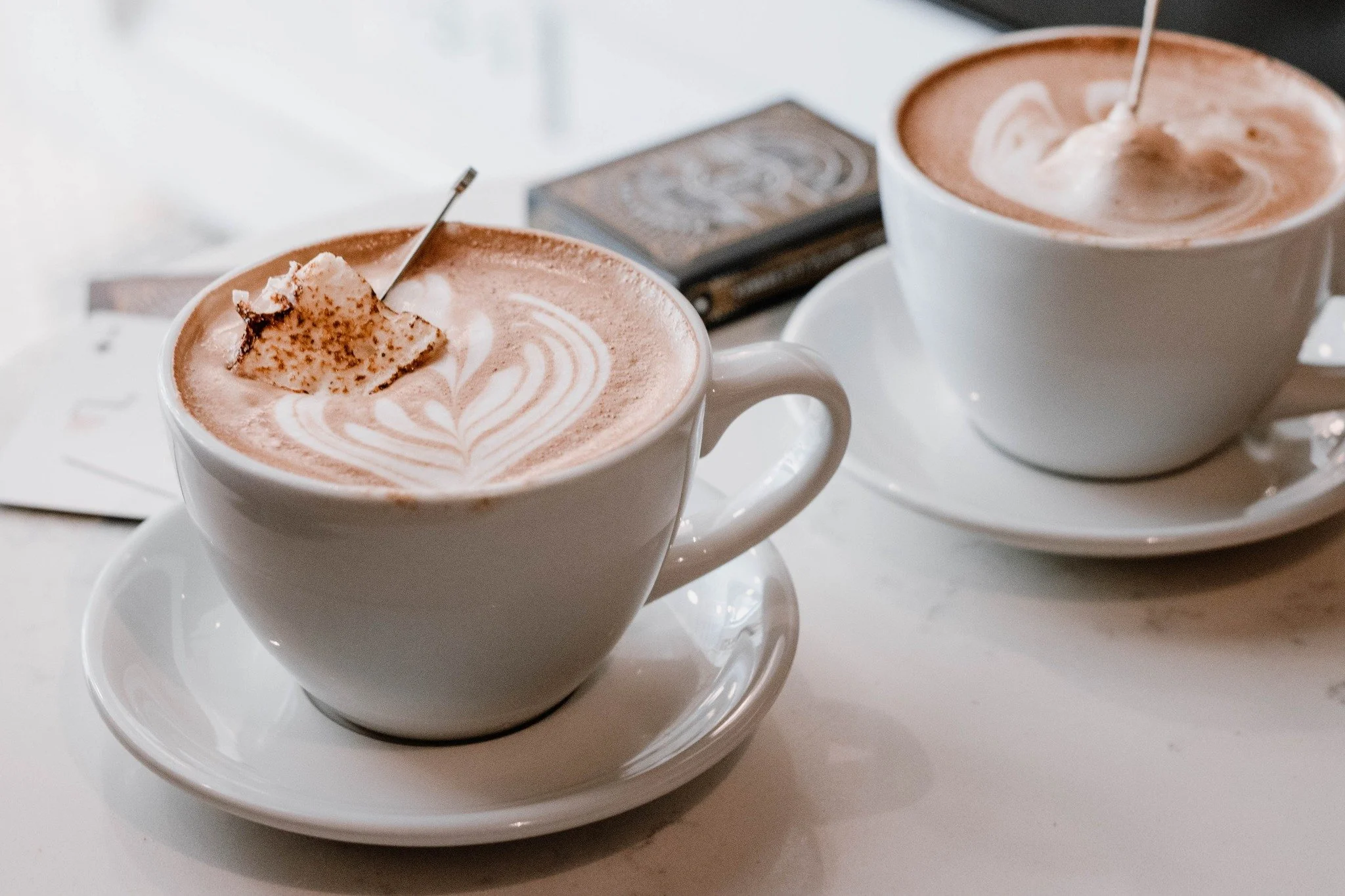 Two white cups of hot chocolate with latte art, topped with marshmallows and cinnamon, on white saucers, with a small chocolate bar in the background.