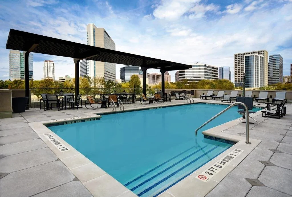 Empty rooftop swimming pool with chairs and tables, city skyline in the background, and a blue sky with some clouds.