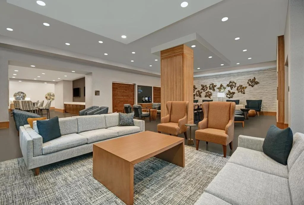 A spacious hotel lobby with modern furniture, including sofas and armchairs, a wooden coffee table, with a background of a bar area and decorative wall art.