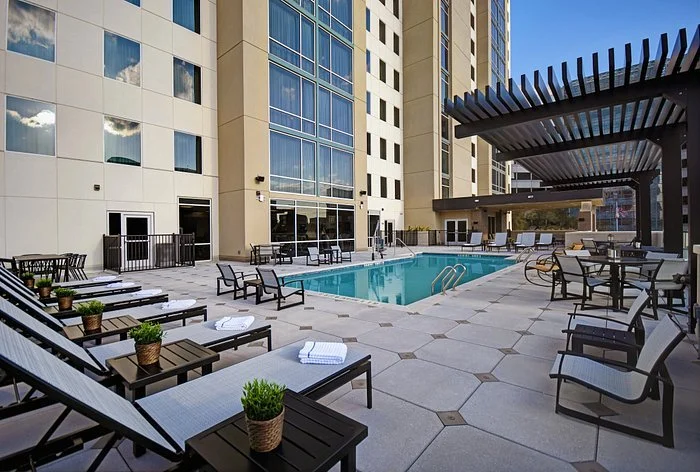 Outdoor hotel pool area with lounge chairs, tables, and potted plants, surrounded by tall modern buildings.