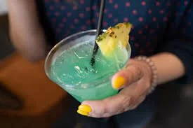 Person holding a blue cocktail garnished with pineapple and a straw.