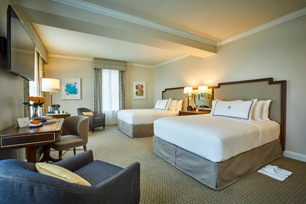 A spacious hotel room with two queen beds, a sitting area with chairs and a small table, a desk with a lamp and a TV, and artwork on the walls.