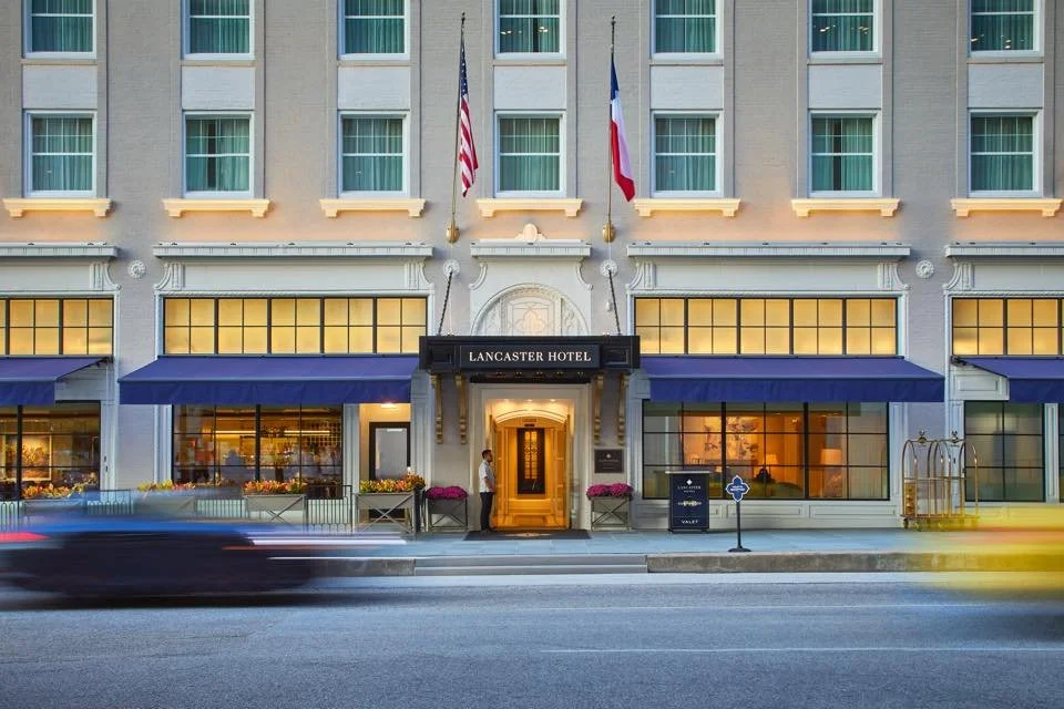 Facade of Lancaster Hotel with American and French flags, blue awnings, and illuminated entrance at dusk.