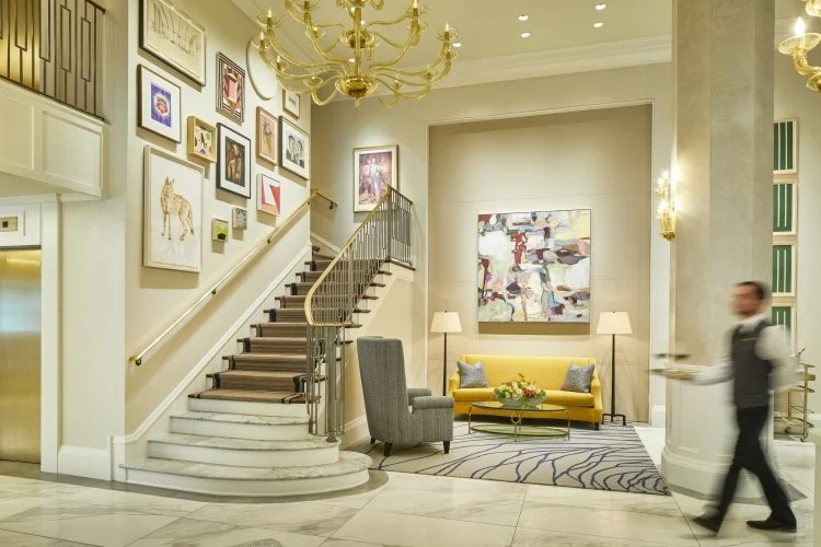Hotel lobby with a staircase, gallery wall, yellow sofa, armchair, abstract painting, chandelier, and a staff member walking by.
