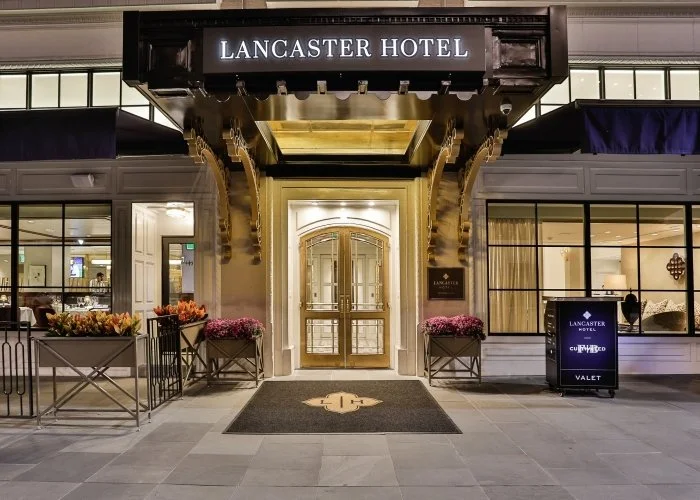 Entrance to Lancaster Hotel with a gold door, black awning, and hanging sign, surrounded by flower planters and a valet sign.