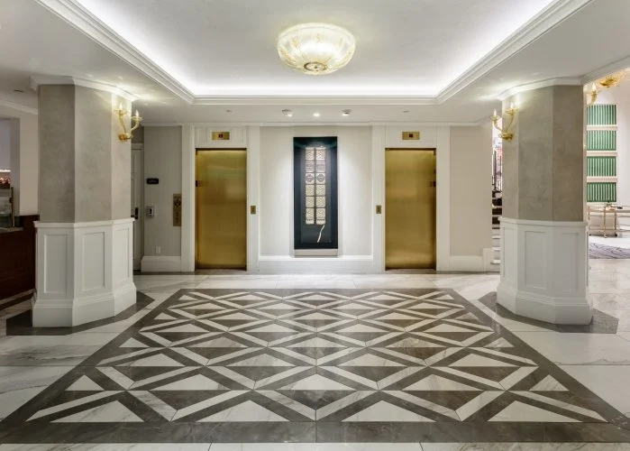 Lobby area with two gold elevators, an ornate chandelier, and decorative wall sconces, featuring a patterned marble floor.