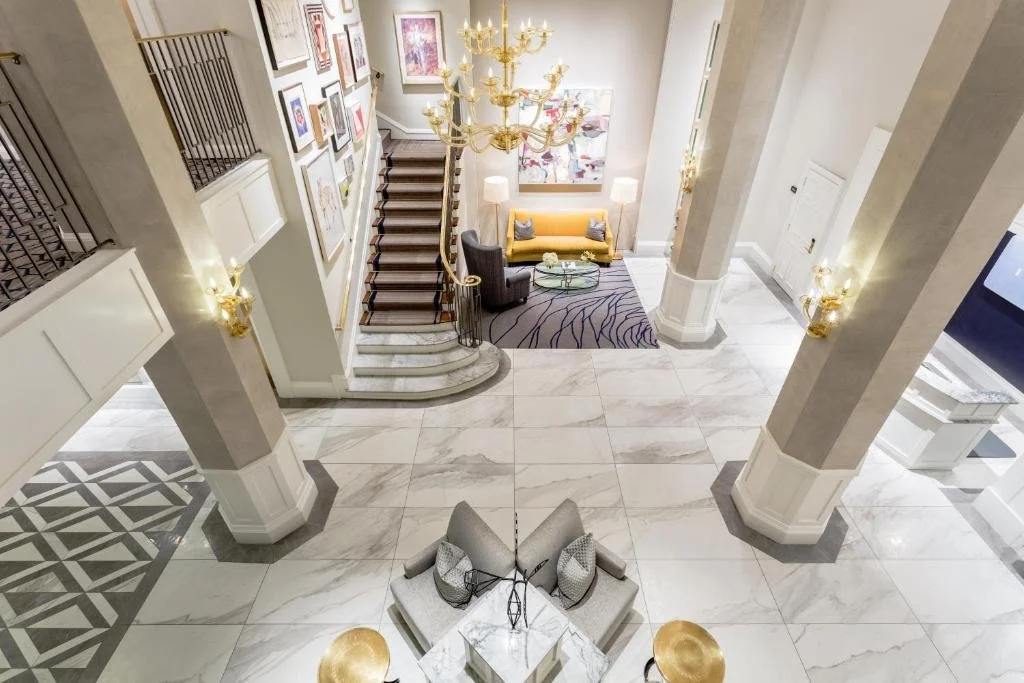 A luxurious hotel lobby with white marble floors, tall columns, a staircase with wooden steps and gold railing, a chandelier, artwork on the wall, a yellow sofa with throw pillows, a purple rug, two white table lamps, and a seating area with armchair