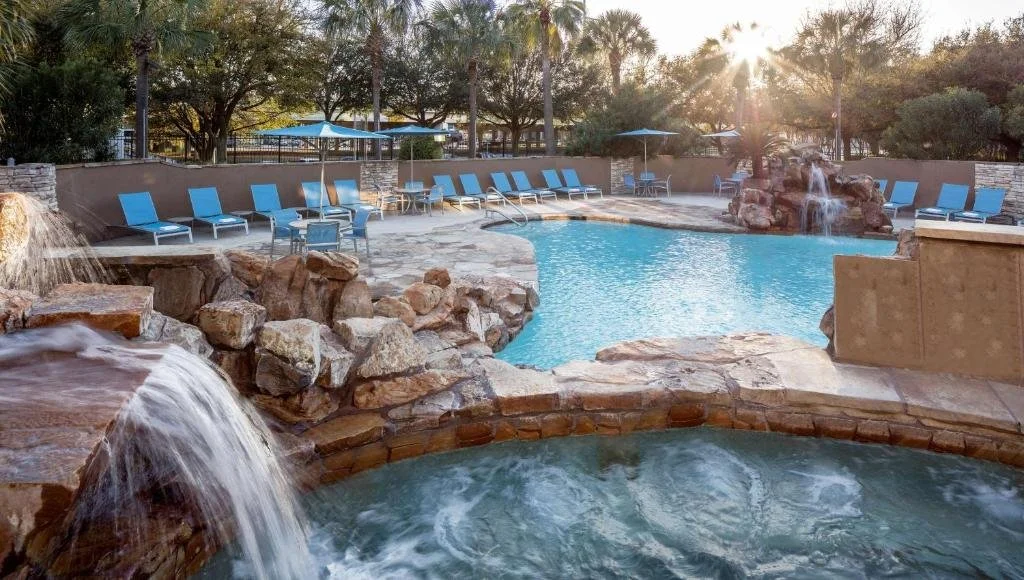 Outdoor swimming pool area with lounge chairs, umbrellas, waterfalls, and rocks, during sunset.