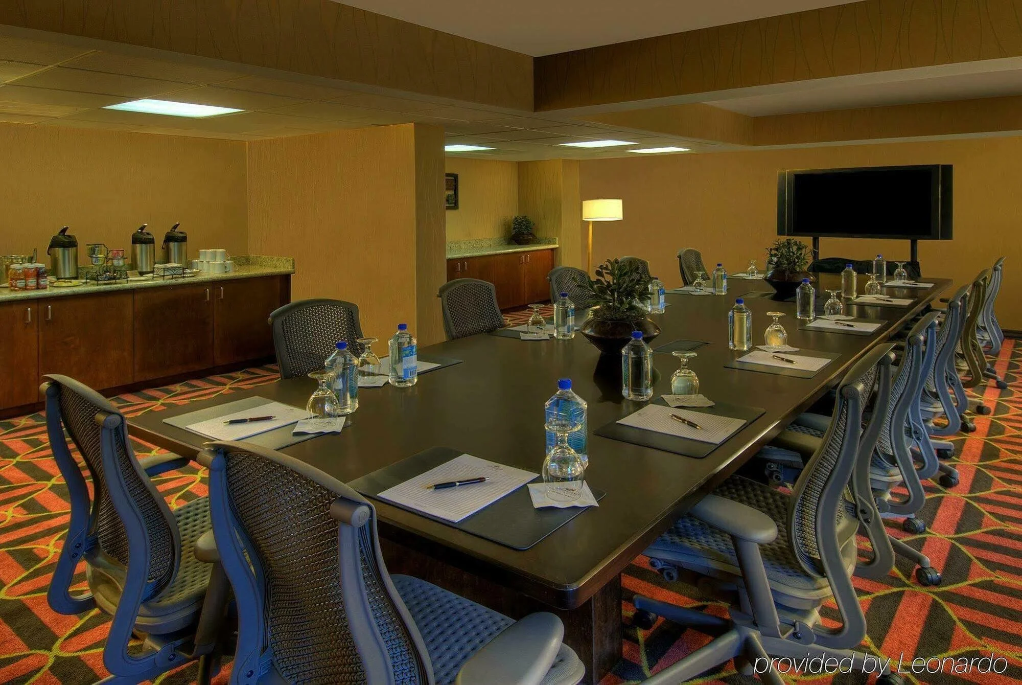 Conference room with a long table set with notepads, pens, glasses, and bottled water. Chairs surround the table, and a large flat-screen TV is mounted on the wall. A coffee station with thermoses and cups is visible in the background, along with a f