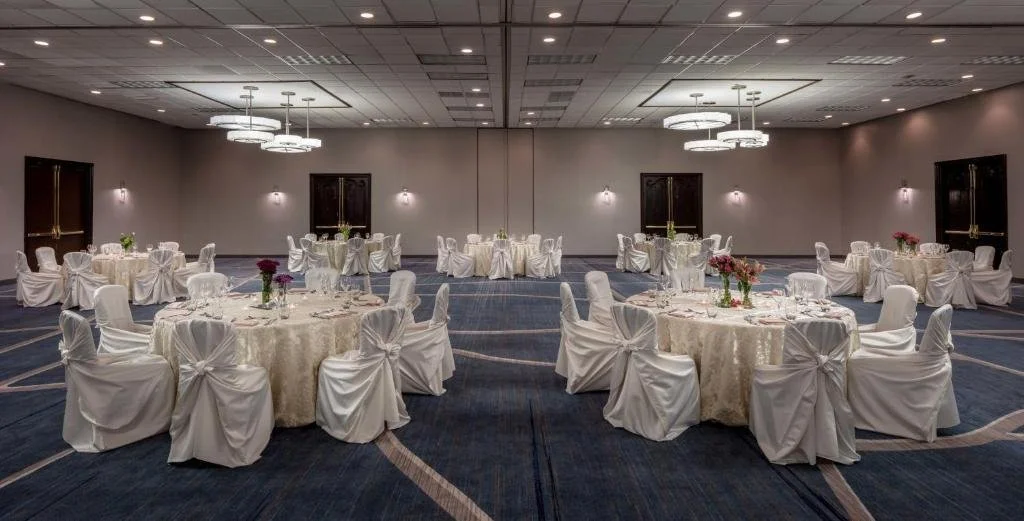 Elegant banquet hall with round tables covered in white tablecloths, adorned with floral centerpieces and surrounded by white chairs with bows, in a neutral-colored room with modern lighting.