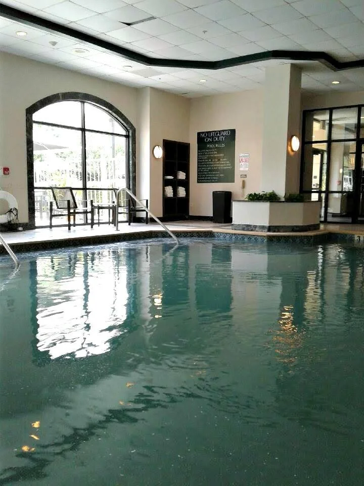 Indoor swimming pool with metal handrails, natural light from large window, and a sign listing pool rules.