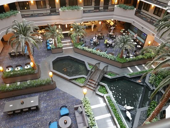 Interior of a multi-story shopping mall or hotel with a central open area, indoor fountain, palm trees, plants, and seating areas, including tables and chairs, with people dining on the upper level.