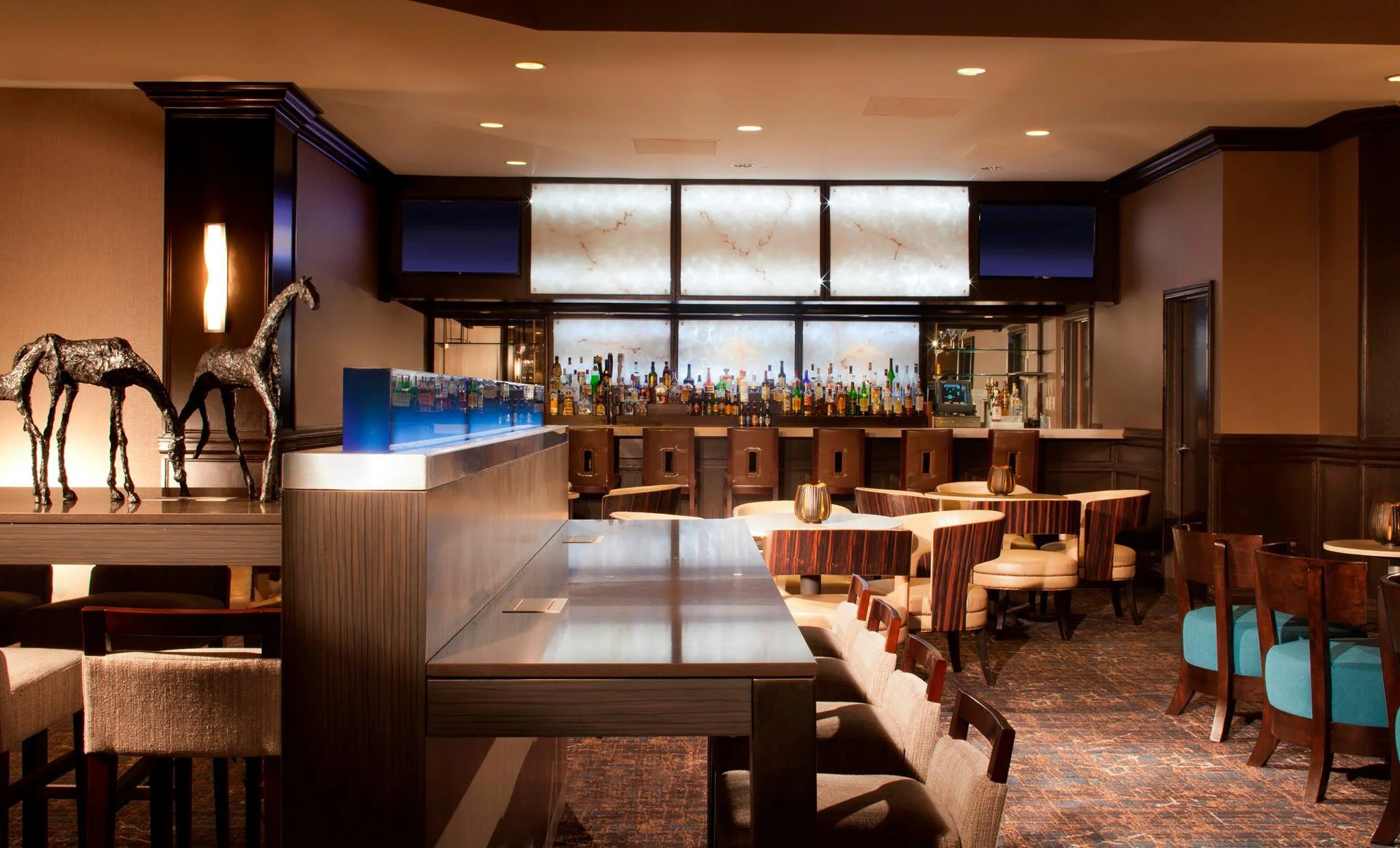 Elegant hotel bar with a variety of liquor bottles behind the counter, modern lighting, and seating arrangements including chairs with blue upholstery and bar stools.