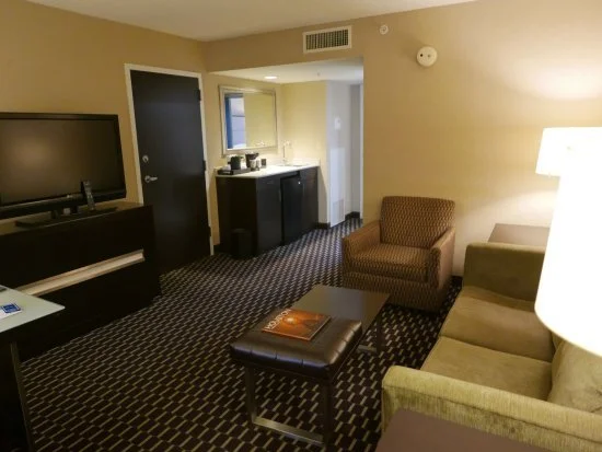 Hotel room with a flat-screen TV, a small sofa, an armchair, a coffee table, a side lamp, a mini refrigerator, and a window.