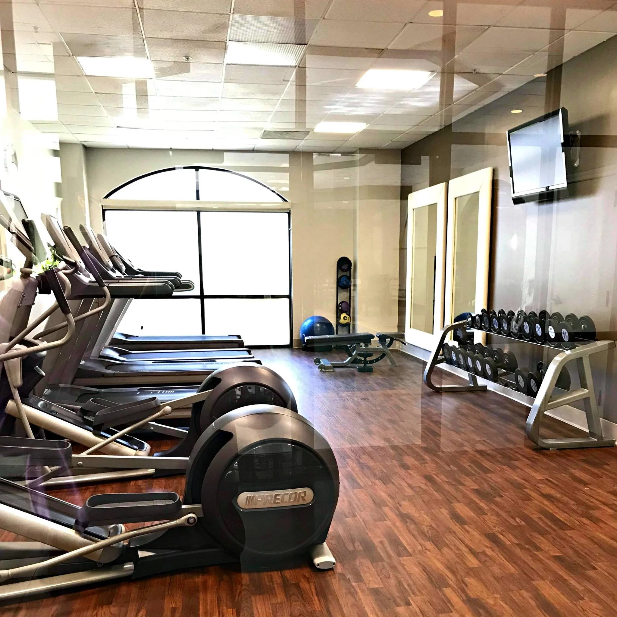 Empty fitness gym room with cardio machines, free weights, mirrors, and a flat-screen TV mounted on the wall.