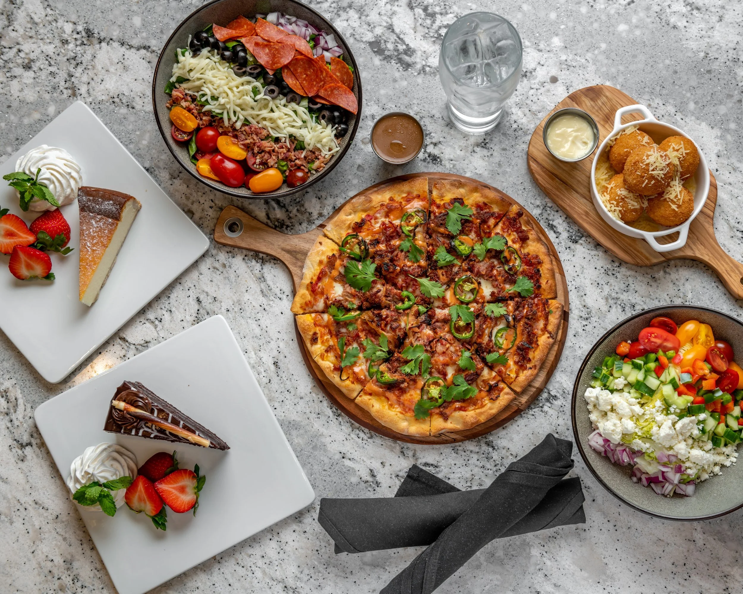 A spread of various foods including a pepperoni and vegetable pizza on a wooden board, two salads with fresh vegetables, a bowl of fried cheese balls with dipping sauce, two slices of strawberry cheesecake, and a fresh fruit and vegetable salad on a 