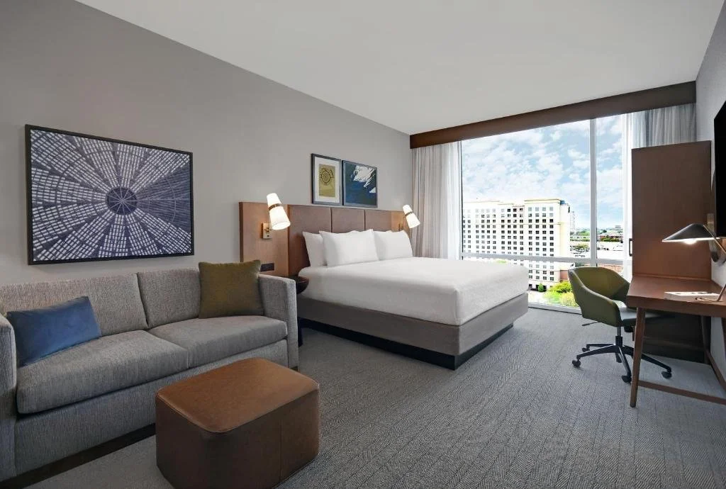 A modern hotel room with a large bed, a gray sofa with pillows, artwork on the wall, a desk with a green chair, and a large window showing a cityscape with a blue sky.