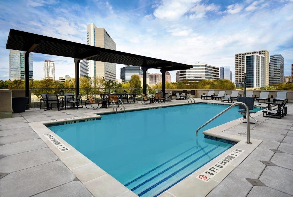 Empty rooftop swimming pool with city skyline in the background, poolside chairs, tables, and a shaded area.